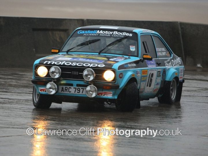 Pistonheads - The image features a vibrant blue rally car in the midst of an auto race on a wet track. The car, likely a Mitsubishi Lancer given its resemblance, is adorned with a variety of decals, including an 'Amazing Grace' sticker prominently displayed on the side. The car's four square headlights are illuminated, cutting through the rain, and the text "RALLYNATION.CO.UR" can be seen on the side of the car, possibly indicating its registration or affiliation with a racing event. The wet surface of the track and the surrounding environment suggest a rainy day suitable for such high-speed pursuits.