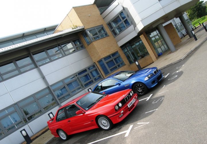 Local photo locations - Page 1 - East Anglia - PistonHeads - This image features a parking area with two sporty cars. The first car is a red coupe with white accents, parked facing the building. The second car is a bright blue convertible, also parked facing the building. Both cars are positioned in alternating spot-markers, indicating that no more vehicles can be parked in the back. The building in the background has a brick facade and large glass windows, suggesting an urban setting. The sky is slightly hazy, but appears to be a clear day with good lighting, accentuating the cars' colors and aggressive designs.