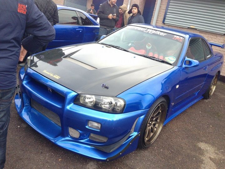 A blue car is parked in front of a building - Pistonheads - The image shows a group of people gathered around a blue sports car, which is a Honda Integra Type R with a widebody kit and additional aftermarket parts. The car is parked on a street next to a building. The car's body is blue and it has a black hood, giving it a distinctive appearance. It also has custom aftermarket parts such as a large rear spoiler and a front bumper with vents. The setting suggests this might be a gathering or event where car enthusiasts come to show off their modified vehicles.