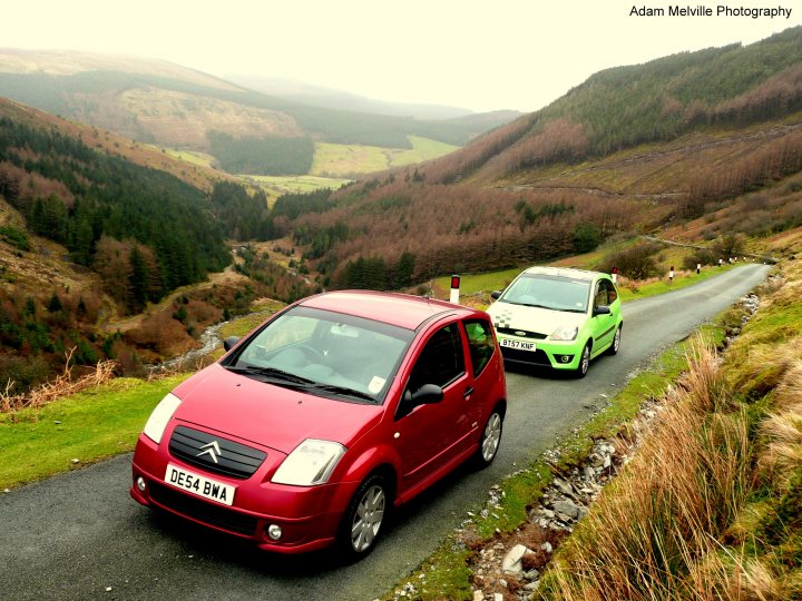 Pistonheads Triangle Evo - The image captures a scenic mountain landscape with a narrow, winding road. Two cars, a red one and a green one, are driving on the road. The red car is closer to the viewer and is positioned on the left side of the road. The road is surrounded by dense trees and grass on the side. In the distance, the expansive mountain range is visible, with the highest peaks shrouded in mist or possibly a growing layer of white frost, suggesting a low temperature environment. The image is watermarked at the top with the words "Adam Meiville Photography."