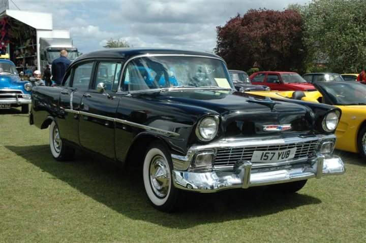 Fins and Chrome - Page 1 - North East - PistonHeads - The image displays an old-fashioned black car, likely from the 1950s, with white-walled tires, prominently facing forward. In the background, there are other vintage cars and trucks on display, suggesting that this scene might be part of a classic vehicle show or gathering. The setting seems to be an outdoor event or field, with vehicles parked on grass, and the sky is overcast. The focus of the image is primarily the black vehicle in the foreground, with the vehicle in focus and the others appearing less sharp due to the wide-angle lense, which can sometimes soften background details.