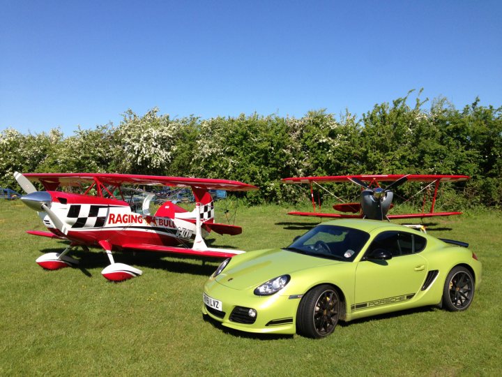 Boxster & Cayman Picture Thread - Page 4 - Boxster/Cayman - PistonHeads - The image captures an outdoor scene of two small aircraft and a modern sports car parked side by side on a grassy field. The aircraft have propellers and are predominantly red with a checkered pattern. The sports car is bright neon green. The sky is clear and blue with small white clouds, suggesting good weather. The green of the field contrasts with the blue of the sky and the red of the aircraft. The arrangement of the objects, one ahead of the other, gives a sense of progression or preparation for a journey or adventure. The vehicles' placement on the grass indicates a possibly casual or informal setting. Overall, the image portrays a visually striking situation where sport and adventure are presented.