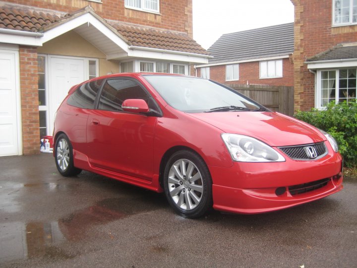 Pistonheads Daventrys Newest Resident - The image features a parked red Honda Jazz on a residential street. The car is positioned at an angle to the camera, allowing its side profile to be fully visible. The vehicle is parked in front of a gray brick house with white trim around the windows and a brick driveway. There is a sidewalk adjacent to the driveway, and a fence can be seen behind the car. The ground appears wet, suggesting recent rain.