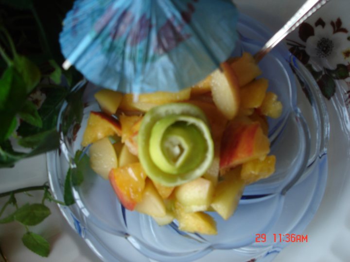 The image shows a bowl of sliced fruit placed on what appears to be a dining table. The bowl contains a variety of fruits, including apple and pineapple. On top of the bowl is an open blue parasol, which adds a vibrant pop of color to the scene. There is also a white table setting visible, along with a silver spoon and a partial view of a plant. The photo is watermarked with the date "29/1/98," indicating it was taken on that specific day.