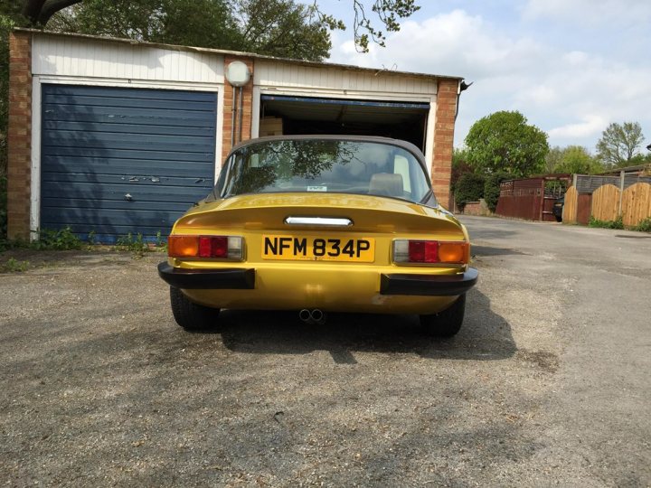 Early TVR Pictures - Page 90 - Classics - PistonHeads - The image depicts a vibrant yellow car parked outside a garage with a wooden door and a large number 834 displayed on the back. The vehicle has the distinguishing features of a classic era car, such as a spare tire mounted under the trunk lid and sideki na. There's also a trailer in the distance, and a solitary tree can be seen in the background on the driveway, suggesting a peaceful suburban setting.