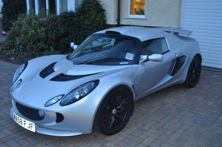 I'm better now - Page 1 - Elise/Exige/Europa/340R - PistonHeads - The image shows a sleek, gray sports car parked on a brick driveway outside a residential building. The car appears to be a two-seater with a convertible top, featuring black rims, a distinctive front grille, and illuminated headlights. The vehicle has a glossy finish and the road behind indicates it's parked next to a curb.