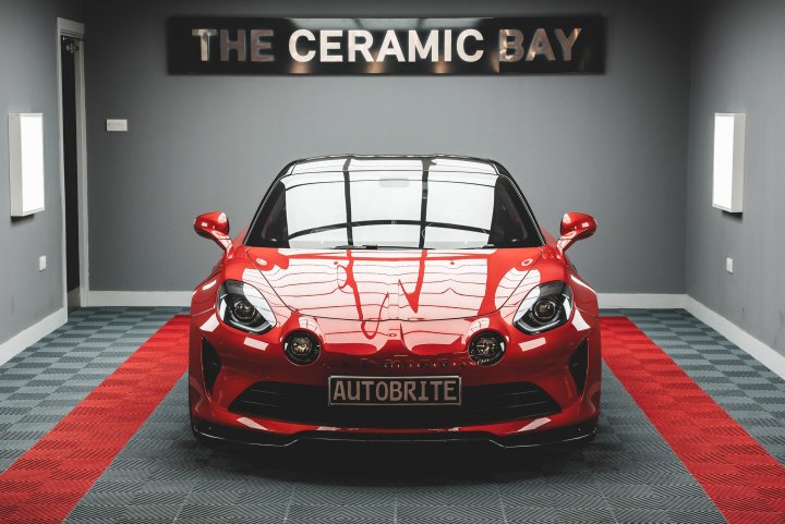 My new Alpine A110S Aero gets detailed by Autobrite Direct  - Page 1 - Alpine - PistonHeads UK - The image showcases a vibrant red sports car parked in a room with a carpeted floor. The vehicle is sleek and shiny, indicating its well-maintained condition. Above the car, there's a sign that reads "THE CERAMIC BAY," suggesting it might be part of an exhibition or event related to ceramics. The room also has red curtains lining the windows in the background, providing a matching backdrop to the car. The overall impression is one of luxury and artistry, possibly hinting at a high-end car showroom or gallery.