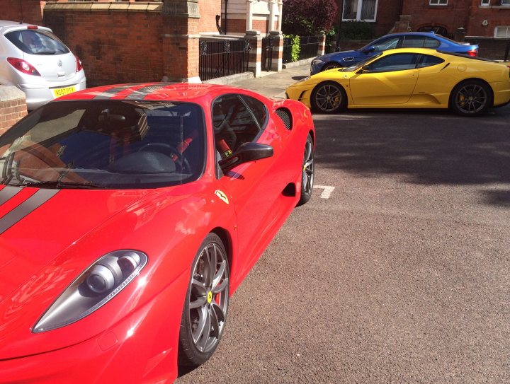 Incoming Scud! - Page 1 - Ferrari V8 - PistonHeads - The image depicts a street scene with two sports cars parked on the pavement. The car on the left is a bright red Ferrari, exhibiting its distinctive design features such as large air vents and a curved windshield. The second car, parked behind the Ferrari, is a sunny yellow Lamborghini, known for its sleek shape and impressive speed capabilities. The background reveals a brick building, suggesting a residential area. A single person can be seen in the scene, partially visible on the left, likely associated with the Ferrari due to proximity.