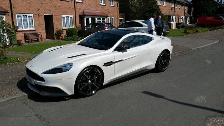 I only went in for a service........ - Page 4 - Aston Martin - PistonHeads - This image captures an urban scene with a white coupe as its central focus. The car is parked on the side of a quiet road, its luxurious design sharply contrasting with the more mundane environment. The coupe is a modern, sleek vehicle with a prominent front grille. The background reveals residential buildings and a truck parked on the grass, suggesting a suburban setting. There are people present, and it appears one person might be about to enter the car. The weather is clear, with the sunshine fully illuminating the surroundings, casting a soft light on the scene.