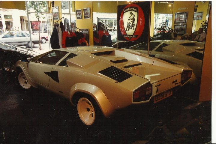 Countach  - Page 13 - Lamborghini Classics - PistonHeads - This image features a golden, retro-style car parked inside a showroom or auto shop. The car has a distinctive white stripe across its side and features a rear spoiler that is prominently visible. The showroom has mirrored walls on both sides of the car, reflecting the vehicle and creating a double impression. There are other cars visible in the background, indicating that this is a place where various cars are displayed. A window in the background shows the view outside of the venue.