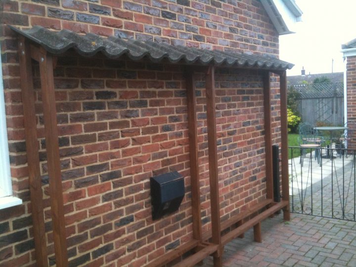 Woodburners Pistonheads Wood - The image shows the outside of a brick building with a flat roof. Attached to the side of the building is a wooden structure that appears to be a small porch or awning. Underneath this overhang, there is a black mailbox attached to the wall of the building. The building is located on a residential street, and the brick wall and woodwork suggest an urban environment. In the background, there is a wrought iron fence and a sign indicating a "no parking" zone.