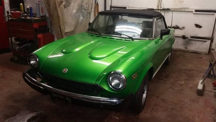 A blue car parked in a parking lot - Pistonheads - In the image, I see a vintage car with a vibrant green body and a shiny hood. The car is parked facing the camera, positioned on what appears to be a maintenance or shop floor. The front of the vehicle is clearly visible, with the round headlights and a distinctive grille, which suggests it might be an early to mid-20th-century model. In the background, there are shelves with wheels and other parts, indicating the setting is a garage or workshop. The floor is dirty, which is typical of a workspace. There are various tools and maybe some other equipment scattered around, which are related to car maintenance or repair.