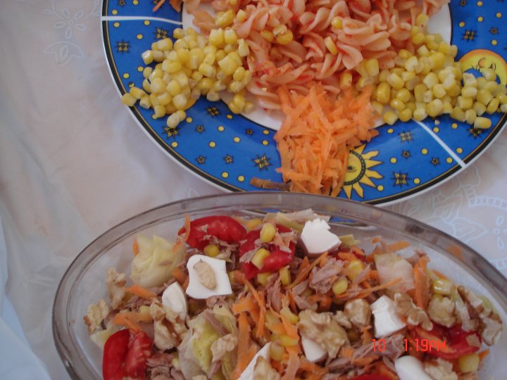 The image presents a vibrant meal scene. On the left side of the table, there's a glass bowl filled with a colorful salad of chicken, corn, carrots, and red peppers. The salad appears fresh and healthy, with the chicken and vegetables providing a beautiful contrast of whites and greens. 

On the right side of the table, there's a white bowl containing a pasta dish. The pasta looks like it might be penne given its tubular shape, and it's smothered in a creamy sauce. The dish is garnished with bits of corn and carrot, adding to its visual appeal and possibly, its taste. 

In the background, there's a blue and white plate holding dessert. The dessert consists of sliced pie under a generous dollop of whipped cream, which stands out with its creamy texture. The dessert's colors and textures provide a pleasing contrast to the savory dishes. 

Overall, the image suggests a well-balanced, appetizing meal served on a white tablecloth. The dessert, salad, and pasta dish are all neatly arranged on their respective plates, ready