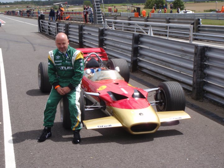 Pistonheads Snetterton - In the image, a man is sitting on the side of a bright red race car on the track. He is dressed in a green and yellow racing suit. The sleek red and gold race car has black wheels. A metal barrier surrounds the track, separating the racing area from spectators. The race car appears to be on display, perhaps for pre-race photographs or interviews.