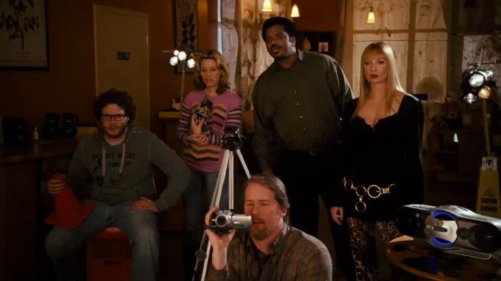 In this image, we see a scene from a television show or movie. It features five characters in what appears to be a retro setting, possibly a set designed to represent the 1980s. The characters are assembled around a television camera crew, suggesting that they could be actors or hosts preparing for a broadcast. The camera operator is prominently featured in the foreground, framing the other characters in the background. The overall atmosphere is that of a taped or live show, evoking a sense of anticipation among the characters.