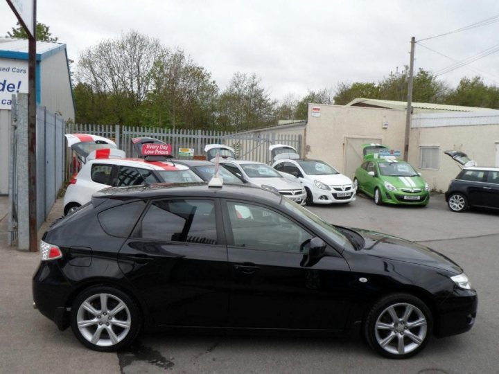 Impreza as a first car - Page 1 - Subaru - PistonHeads - The image shows a parking lot with various cars parked in different directions. In the foreground, a black sedan is prominently displayed, occupying a large part of the image. A few other vehicles, including smaller cars and a hatchback, are also visible in the background. To the side of the lot, there is a small building with a sign displaying the words "STEP OUT" and "WED Car hire." The sky is overcast and the lot seems relatively empty.