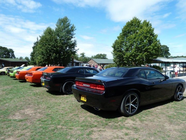 Challengers at Brooklands 09/08/15 Photos (please add) - Page 1 - Yank Motors - PistonHeads - The image depicts a line of cars parked side by side in what appears to be a spacious outdoor area. The cars are designed with dual rear wheels, a feature commonly associated with certain performance vehicle models. The car closest to the viewer is a dark blue model with a license plate that is not fully visible due to its angle and the lens flare that partly obscures it. Behind this car, other similarly styled models in various colors can be seen, including one in a vibrant hue of orange and another closer in tone with blue. There are people visible in the vicinity of the cars, contributing to a social or event atmosphere. The background features a mix of sky, trees, and structures that do not take the focus away from the cars. Overall, the image captures a moment of gathering or display of these distinctive vehicles.