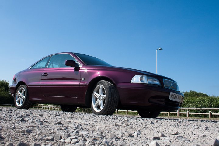 Who's got what in the South West - Page 1 - South West - PistonHeads - The image showcases a vibrant purple coupe parked on a gravel road with a grayish ground surface. A fence is visible in the background, suggesting a rural or semi-rural setting. The sky is a bright blue, indicating a clear and sunny day. The car, with its shiny paint and shiny exterior, is the central focus of the image, displaying a sense of depth as it is captured at a diagonal angle.