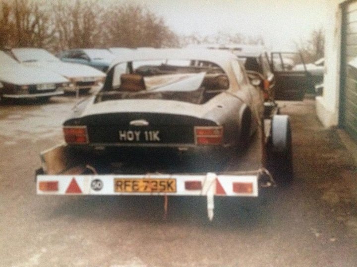 HOY 11K 4wd TVR se restoration project - Page 1 - General TVR Stuff & Gossip - PistonHeads - This is an outdoor image showing a black car with an open trunk that appears to have been dumped illegally at the side of a road. The vehicle is parked on a gravel-covered area, and there are other cars in the background, suggesting this might be a parking lot or a street with parked vehicles. The car has a white license plate frame. The sky above the scene is overcast.