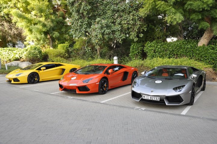 Pistonheads - The image depicts a scene in a parking lot where three luxury sports cars are parked. The yellow car is on the left, followed by the red car with a unique design, and positioned next to it is the silver one. The cars appear to be new, and their bright colors against the gray concrete stand out prominently. A lush backdrop of green trees and shrubs enhances the luxurious ambiance of the image.