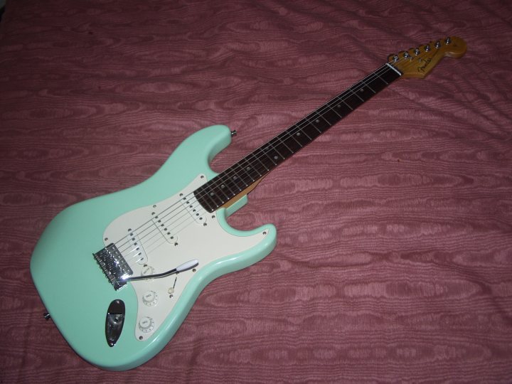 Lets look at our guitars thread. - Page 177 - Music - PistonHeads - The image shows a vibrant turquoise electric guitar placed against a plum-colored fabric backdrop. The guitar is a vintage white Stratocaster model, known for its distinctive white color, three-pickup configuration, and pickup selector switch. The guitar is detailed, with a pickguard and various body contours, reflecting the iconic design of this popular electric guitar. It is also equipped with metal pickups and six tuning pegs.