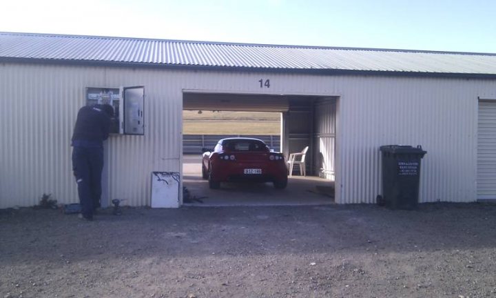 A man standing next to a parked truck - Pistonheads - This image shows a small white shed with a garage door ajar, revealing a red car parked inside. The car is oriented diagonally, with its front wheel facing the closed end of the shed. To the left of the shed, a tall person is standing beside a trash tote, reaching into the shed. In the background, the vast expanse of a desert landscape stretches out. The sky above is clear, suggesting a bright and sunny day. There is a sense of solitude and privacy conveyed by the scene's informal composition.