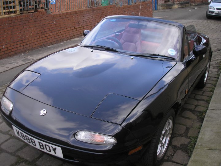 Pistonheads Pricing - The image shows a black sports car parked on the right side of a brick road. The car's convertible top is closed. Above, clear blue skies contrast with the red brick buildings lining the street. There is another vehicle in the far background, suggesting the car is parked in a public parking space. The car's badge is visible, but the specific make and model are not identifiable from this perspective.