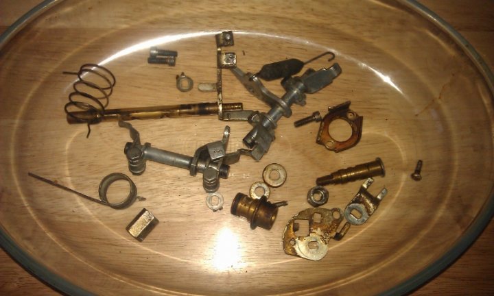 A wooden table with a clock on it - Pistonheads - The image displays a collection of mechanical parts laid out on a wooden surface, likely for repair or maintenance purposes. The items include small metal components, gears, and various other parts with visible wear and rust, indicating they have been used or are old. Notably, there is a rusty metal spring which might be part of a click pendulum from a clock, as well as other devices. Most of the objects are situated at the center of the plate, and they are placed in such a way that it is easy to identify them. Overall, the setting suggests a tinkerer or someone working on mechanical devices.