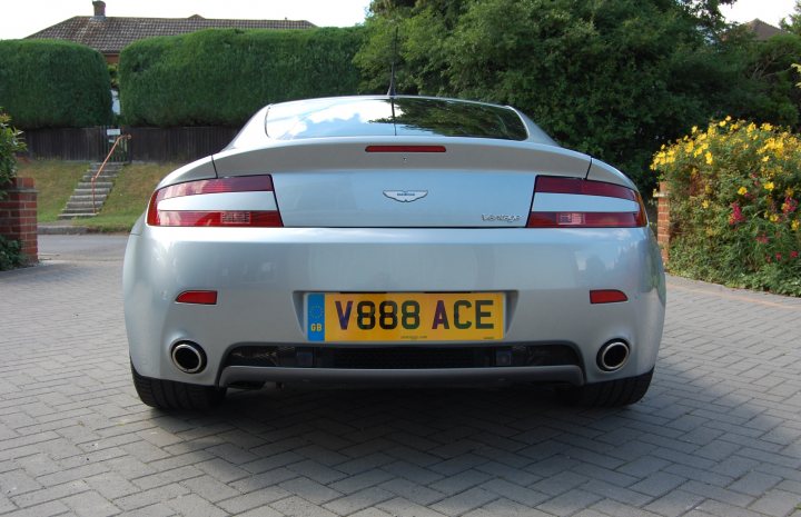 number plates - Page 1 - Aston Martin - PistonHeads - The image features a close-up view of the back of a silver sports car parked outdoors. Its rear license plate reads "V88 ACE." The car appears sleek and well-maintained, with the prominent Ace logo on the tailgate. Beneath the car, a cobblestone driveway extends into the image, and lush green trees and a manicured lawn in the background suggest a residential setting. The sky is overcast, and some flowers can be seen in the distance near what seems to be a wooden fence or a low wall made of similar materials.