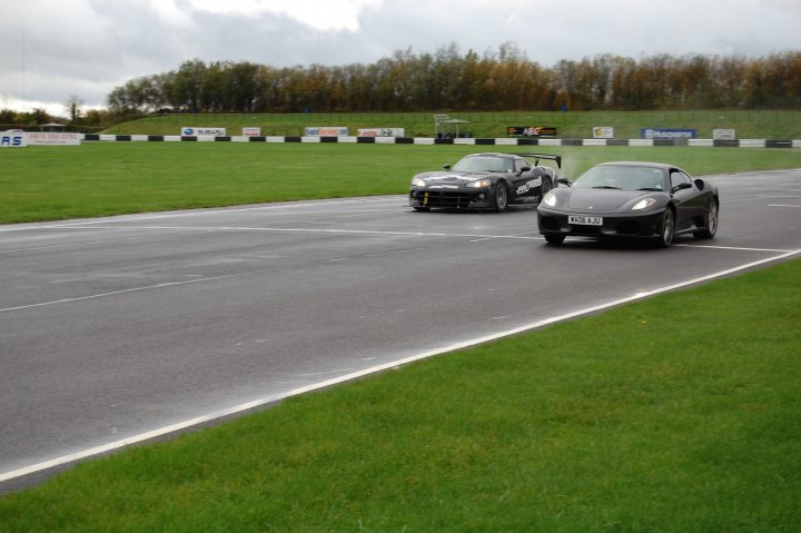 Day Association Stroke Combe Castle Pistonheads Supercar - This image captures an exciting moment on a race track. Two sleek, black sports cars are racing head-to-head, their rivalry palpable as they skid around a crowded track. The track itself is an asphalt flanked by grass on both sides, adding a touch of tranquil greenery to the high-speed action. The background is filled with a hilly landscape, providing a scenic backdrop to this thrilling scene.