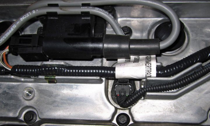 Couple of K series eng sensor Q's - Page 1 - Caterham - PistonHeads - The image shows a close-up view of an engine bay, focusing on a complex tangle of wires and cables. Among the components, there is a black plastic connector and a metal interface that suggests the viewer is looking at the car's electrical system. The wires are mostly black, contrasting with a metallic grey engine cover that forms the backdrop. The overall impression is one of order amidst complexity, as the layout is systematic yet intricate.