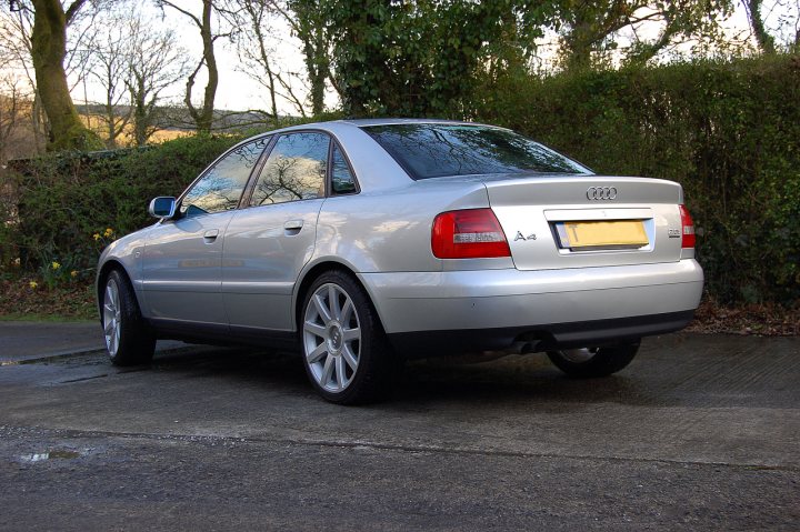 A car is parked on the side of the road - Pistonheads - The image depicts a silver Audi sedan parked on a driveway, adjacent to a low wall. The car has a European-style license plate, suggesting it might be registered in the United Kingdom or similar countries. The vehicle is positioned at a slight angle to the viewer, with the front door on the right. The surrounding area has lush greenery, indicating a suburban setting, and the sky above appears overcast.