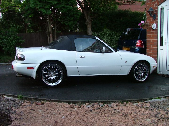 Springs Pistonheads Trim Kyb - The image shows a classic white two-seater sports car that appears to be a part of a private residential setting. The vehicle has its top down, revealing a convertible design. The car is parked on what looks like gravel and is adjacent to a paved driveway. In the background, there is a house with red brick paving and a neatly trimmed hedge. The style of the photo seems candid, capturing the car from a slightly elevated angle in a residential environment.