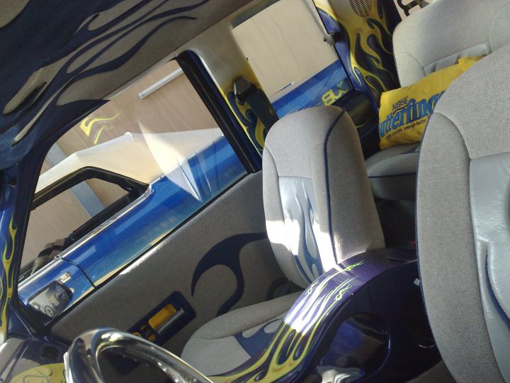 Truck Pistonheads Chevy - The image shows the interior of a car, focusing on the driver's side. The upholstery features a distinctive flame print pattern, and a blue car is visible through the car's windows, suggesting the vehicle is parked outside. The driver's seat has a headrest and appears to be part of the car's customized decor, potentially with an orange and yellow affinity. There is also some cargo visible in the car, including what looks like a yellow box and a bag. The overall feel of the car suggests personalization and style.