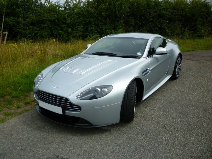 How about an Aston photo thread! - Page 1 - Aston Martin - PistonHeads - The image portrays a luxurious silver Aston Martin sports car parked on a paved surface. The car has a sleek design with a wrap-around windshield and a sloping roofline. Its wheels are large and feature a multi-spoke design. The vehicle is situated in a grassy area with trees in the background, under what appears to be an overcast sky.