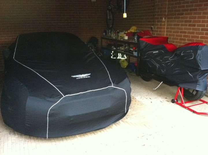 Pistonheads - The image shows a black convertible car parked within an open garage, with its top down, allowing for a clear view of the interior. A black car cover is draped over the hood, secured by straps in an X shape at the corners. There are various tools and items scattered around the garage. Adjacent to the car is a red dolly with a smaller hooded item on it, which is identifiable as a red and black helmet. The garage itself has a red brick wall in the background.