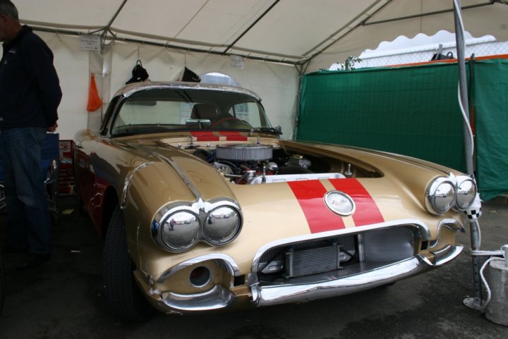 Spotted Earlier Pistonheads - The image shows a vintage muscle car parked underneath an open tent or awning. The car is a pointy model with a prominent hood scoop and an engine visible within it. It features a silver front and is painted in a gold or brass color with a thin red stripe running across its roof. The car is displayed for the public, indicated by its position under the tent and its likely purpose being an exhibit or presentation.