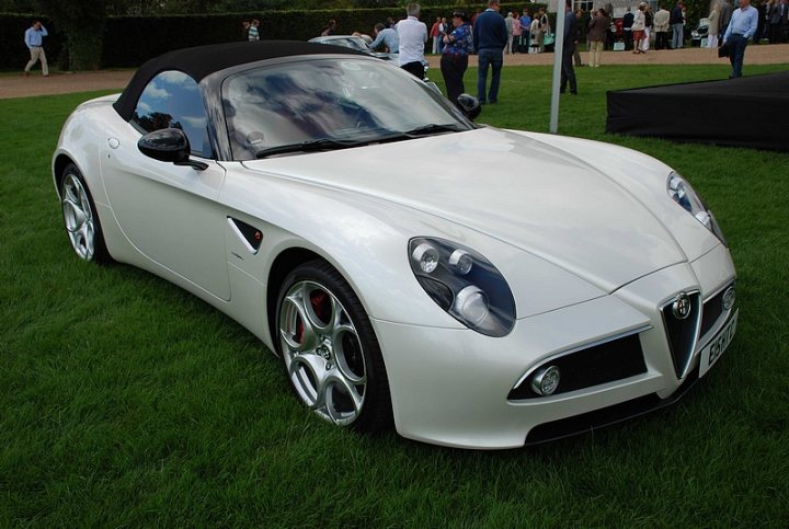 GRRC Lunch Club - Page 1 - Goodwood Events - PistonHeads - The image shows a white, open-top sports car parked on a grassy field. The car has shiny silver wheels and a clean, sleek exterior. It's daytime and the surrounding area appears to be a relatively small outdoor event or gathering, with several people in the background. The car is positioned at an angle to the camera, giving a view of its side and front.