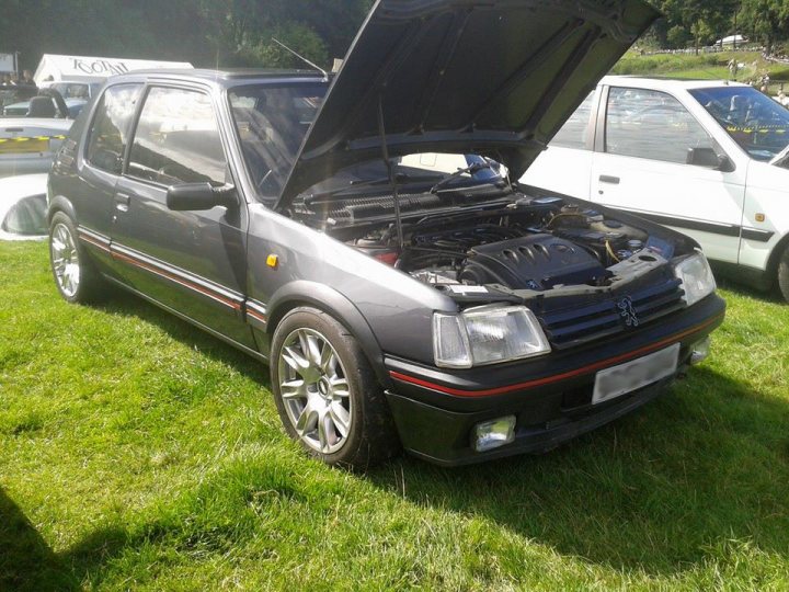 Lets see your Pug! - Page 1 - French Bred - PistonHeads - The image showcases an outdoor scene featuring a black car with its hood up. The car is parked on a grassy area, appearing quite aged or modified, given its pop-up hood and a visible engine block. In the background, another vehicle and a signpost are partially visible.