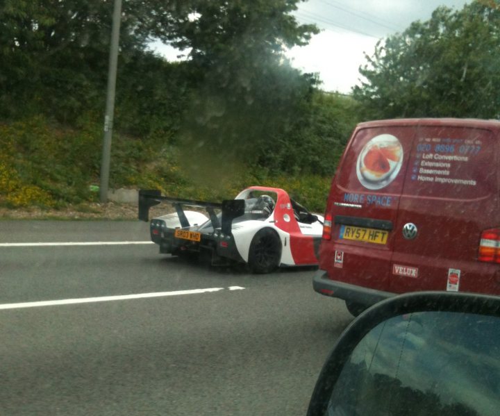 M1 today - Page 1 - Radical - PistonHeads - The image shows a dynamic highway scene, where the car is close to the camera, partially obscured by it. A red Volkswagen van is seen in the foreground, driving behind the low-slung, racecar-like vehicle. The vehicle in front has a large fixed wing and a rear wing, indicative of a race car. The highway is bordered by vegetation, and there's another car further back on the street. The image captures the race car in motion, creating a sense of speed and competition.