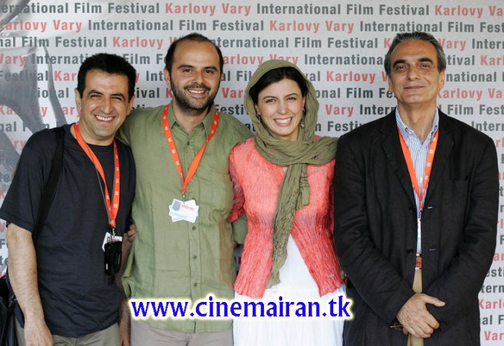 The image is a color photograph of four individuals posing for the camera. They appear to be at a film festival or event, as suggested by the repeated text in the background that includes words like "Film Festival," "Karolyi," and "Various events" in multiple languages. The group includes two men with badges around their necks and one woman. The individuals are dressed in a mix of formal and casual attire, with the man on the left wearing a black shirt and the woman with a bright pink blouse. The man on the right is in a jacket and tie. The atmosphere is cheerful, with smiles on their faces. The background wall is white with text elements in various colors and languages, including English, Spanish, and Italian among others. The overall setting conveys a sense of international cultural exchange.