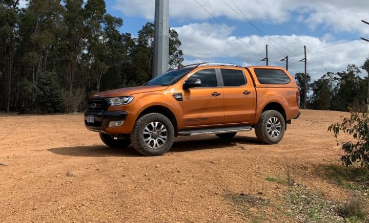 Cooper AT3 4S good on ranger wildtrack - 265/65r18? - Page 1 - Off Road - PistonHeads - The image features a large, orange SUV parked on a dirt surface near a grassy area. The vehicle is the central focus of the image and appears to be in good condition. In the background, there's a structure resembling a water tower, painted white with some visible rust, suggesting an older or abandoned infrastructure element. The sky above is partly cloudy. The overall setting seems to be rural or semi-rural, given the open space and the natural surroundings.