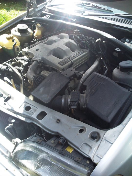 Cortina Pistonheads Ford - The image shows the undercarriage of a vehicle, focusing on the engine compartment. A hood is slightly opened, revealing the engine bay that contains various mechanical components. There are several replacement parts scattered around, suggesting maintenance or repair work. The master cylinder brake booster is prominent, located beneath the engine. Sunlight is streaming onto the engine, casting light on the metal surfaces and giving a clear view of the components. The engine displays a visible brand logo, adding to the identification of the vehicle.