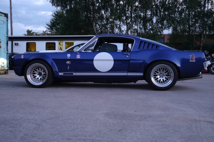 The GT Cafe - the ace cafe of the north - Aug meet - Page 1 - North West - PistonHeads - The image showcases a classic blue Mustang car parked on a grey concrete surface. The car's hood features a distinctive white oval with the "CA" initials at the top center, suggesting it may be a California-registered vehicle, and the new cars sign visible. The Mustang is adorned with red accents and has a black grill, adding to its sleek design. A white star on a blue background is painted on the passenger door, though without more context, the significance of this mark is unclear. The car is positioned in front of a backdrop of other vehicles and buildings, partially obscuring the immediate surroundings but indicating a busy or populated area.