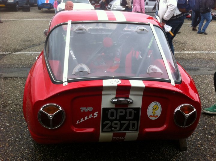 Early TVR Pictures - Page 10 - Classics - PistonHeads - This image features a **red vintage car** sitting in a parking lot. The car has a **white stripe** running along the center of its hood and a **black stripe** on the roof, adding to its vintage charm. It's parked amidst other vehicles, indicating a public parking area. Surrounding the car is a group of people, contributing to the bustling atmosphere of the scene.