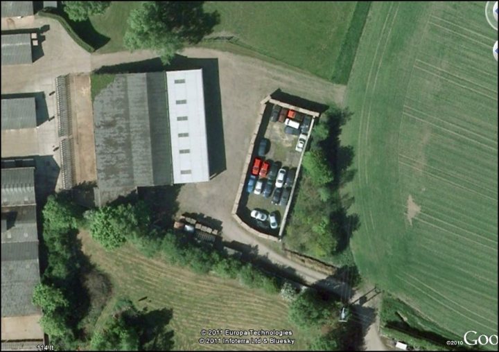 Stolen Pistonheads Hideout - The image depicts an aerial view of a car dealership with a variety of vehicles on display. The dealership appears to be on the outskirts of an urban or suburban area, bordered by a mix of commercial and industrial structures. The vehicular parcel is characterized by several individual car areas, with cars showcased to potential buyers. A prominent feature of the scene is the presence of cows grazing in an adjacent field. The dealership itself has a large rooftop area which adds to the uniqueness of the location. The surrounding area includes other buildings and the layout suggests the presence of multiple dealer operations with a shared perimeter road.