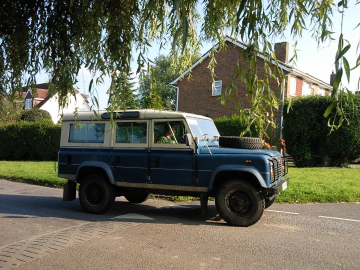 COOL CLASSIC CAR SPOTTERS POST!!! Vol 2 - Page 86 - Classic Cars and Yesterday's Heroes - PistonHeads - This image captures a moment on a city street, where a blue Land Rover is stationed at the bottom of a slight incline. The car is in the foreground, occupying the right side of the street. Behind the Land Rover, a brick house is visible, its green lawn providing a contrast to the urban setting. On either side of the house, there are leafless trees, perhaps indicating a season of change. The street itself appears to be quiet, with no other vehicles or pedestrians in sight.