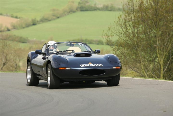 Pistonheads Design Asymmetric - The image captures a moment on a winding road, where a black sports car is in motion. The car is driving on the right side of the road, near a tree-filled embankment. The person inside the car, presumably the driver, can be seen wearing a helmet, adding to the sense of speed. The scene suggests an adventurous drive, with the vehicle leaning slightly due to the bend in the road.