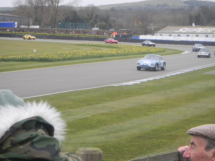 Early TVR Pictures - Page 126 - Classics - PistonHeads - The image showcases a thrilling scene of a race taking place on a narrow track nestled within an open field, encircled by spectators scattered along the perimeter. The track is lined by a blue fence and is perfectly curved, adding to the excitement and anticipation of the race. Several cars are captured in the midst of their sprint, with one particularly close to the finishing line, which is marked by a white line. In the foreground, an individual is seen leaning over a rail, their attention riveted on the unfolding race.