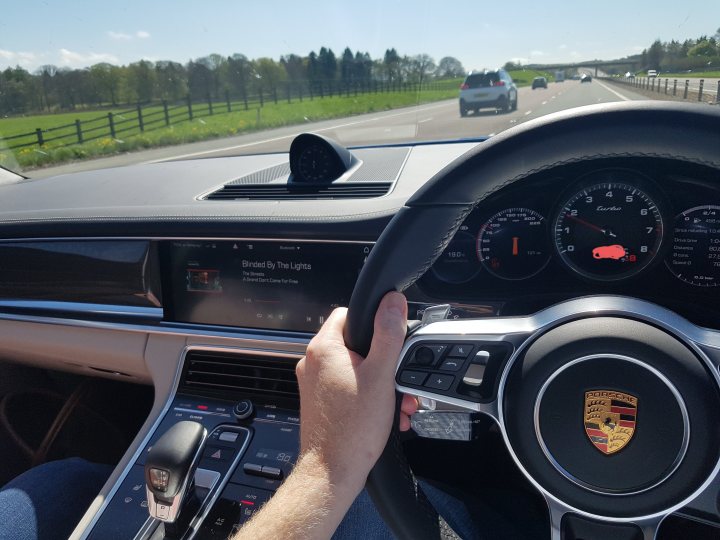 Pics of your Fast Estate... - Page 64 - General Gassing - PistonHeads UK - The image shows a person driving a luxury car, presumably a Porsche, based on the visible logo. The driver is wearing a dark shirt and appears to be focused on the road ahead. The interior of the car is visible, with a dashboard displaying various gauges and indicators. There's a steering wheel with multiple controls in front of the driver. Through the windshield, one can see an outdoor scene with trees and a clear blue sky, suggesting good weather. A highway or main road is visible, indicating that the car is in motion on a paved public road.